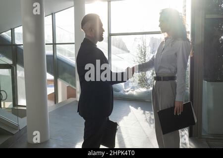 Due giovani e fiduciosi partner commerciali in abbigliamento formalwear che agita le mani mentre si trovano di fronte l'uno all'altro all'interno di un moderno ufficio Foto Stock