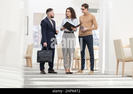 Giovane ed elegante manager femminile con cartella aperta che guarda uno dei colleghi maschi durante la discussione delle strategie all'incontro in ufficio Foto Stock