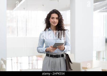 Giovane donna caucasica designer o manager con tablet in piedi di fronte alla fotocamera in grande ufficio moderno con interni bianchi Foto Stock
