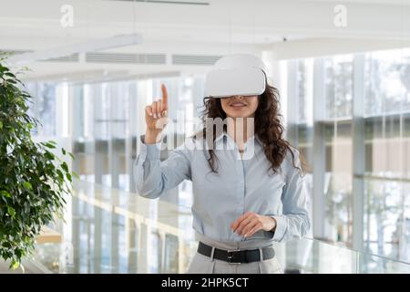 Felice giovane sviluppatore di videogame contemporaneo in visore vr che punta allo schermo virtuale mentre fa la presentazione di nuovo spazio high tech Foto Stock