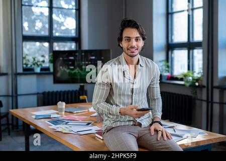 Giovane uomo d'affari di successo in elegante casualwear seduto sul tavolo con carte finanziarie e guardando la macchina fotografica in sala riunioni Foto Stock