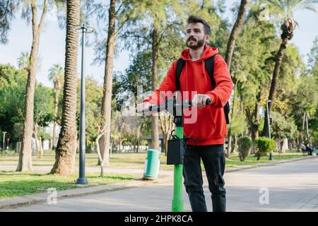 Giovane uomo con zaino in sella a uno scooter elettrico in un paesaggio urbano. Noleggio bici. Foto Stock