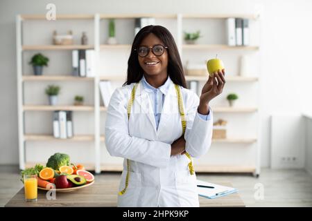 Allegro consulente di nutrizione femminile afroamericana con mela e metro a nastro che lavora in clinica e sorridendo alla macchina fotografica Foto Stock