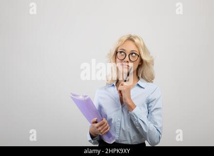 Giovane donna d'affari sognante che tiene cartella con documenti, pensando e guardando da parte lo spazio di copia, sfondo chiaro Foto Stock