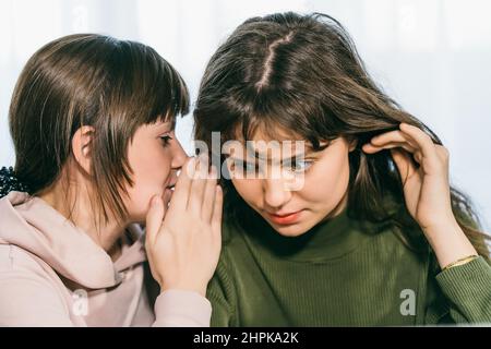 Giovane donna carina sussurrando segreto su orecchio alla sua amica femmina. Comunicazione e ragazza sorpresa da gossip. Foto Stock