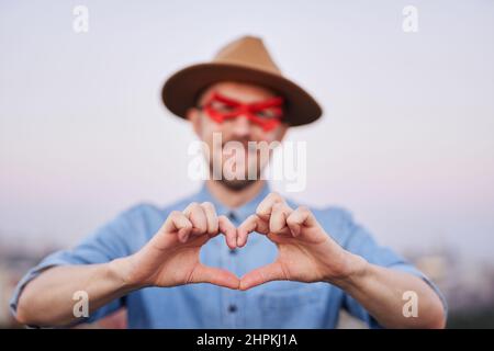 Attraente maschio caucasico in maschera occhi supereroe rosso e maglietta jeans in posa di fronte alla macchina fotografica mostrando amore o gesto di cuore all'aperto al tramonto con vista urbana. Immagine di alta qualità Foto Stock