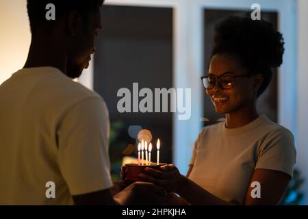 Giovane coppia allegra nera che tiene piccola torta fatta in casa mentre festeggia l'anniversario a casa Foto Stock