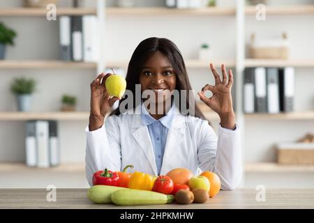 Giovane dietista nero che tiene la mela, mostrando il gesto giusto, seduto alla scrivania con frutta e verdura, sorridendo alla macchina fotografica Foto Stock