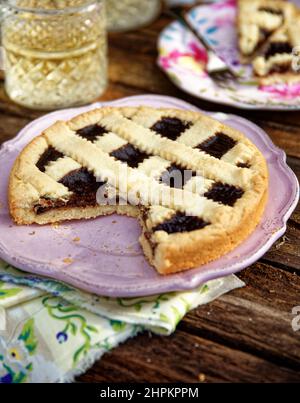 Torta di pasta alla crema di cioccolato servita in un piatto rosa su tavola di legno e tovagliolo verde, fette di torta in campo su un piatto fiorito, due bicchieri di col Foto Stock