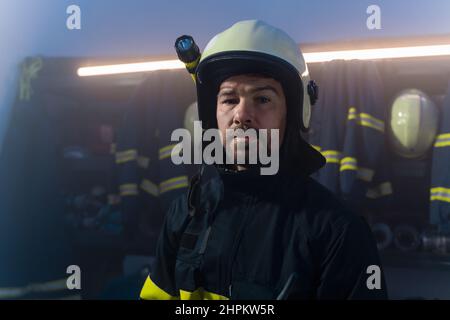 Vigile del fuoco maturo che si prepara all'azione nella stazione dei vigili del fuoco di notte, guardando la macchina fotografica. Foto Stock