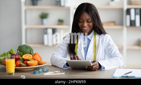 Giovane ragazza blck femminile dietista che lavora con il computer tablet, dando consulenza online o la creazione di dieta piano presso l'ufficio medico, copia spazio, panorama. Nu Foto Stock