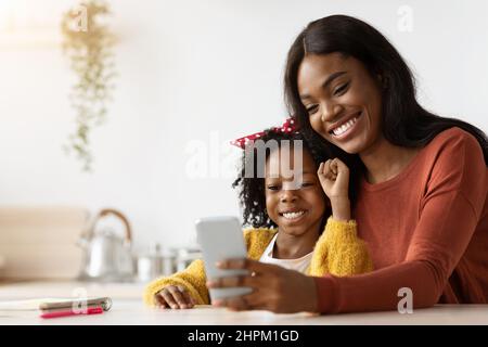 Allegro giovane madre nera e figlia rilassarsi con smartphone insieme Foto Stock