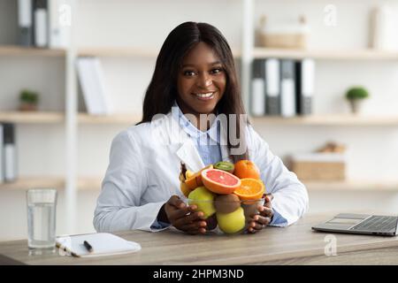 Ritratto di nero femminile nutrizionista seduta al posto di lavoro con ciotola di frutta fresca, lavorando alla clinica di perdita del peso Foto Stock