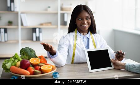 Amichevole afroamericana donna dietista mostrando dieta piano e raccomandare femminile paziente frutta fresca per mangiare Foto Stock