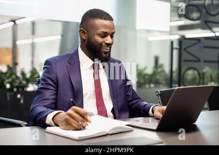 Positivo nero manager digitando sulla tastiera del laptop, prendendo appunti Foto Stock