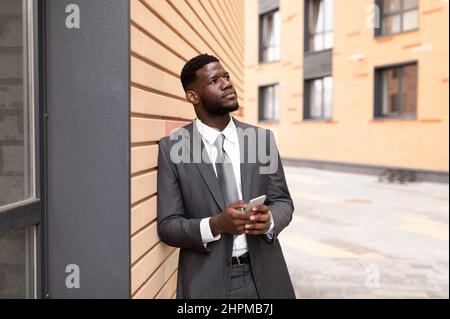 Pensiero strategico. Il pensieroso uomo d'affari afroamericano tiene lo smartphone e guarda via, appoggiato sul muro Foto Stock