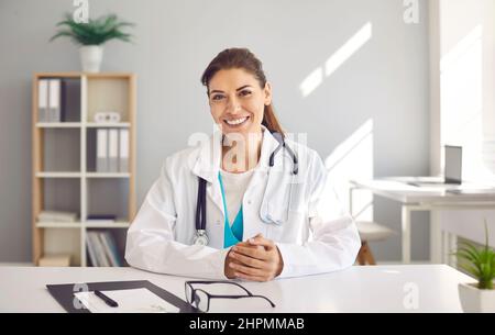 Ritratto di felice sorridente medico femminile in cappotto bianco seduto alla scrivania nel suo ufficio Foto Stock