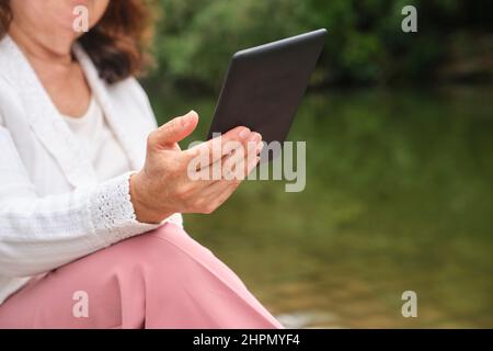 Donna matura irriconoscibile che legge un ebook su un lettore elettronico seduto accanto a un lago in un parco. Concetti: Tecnologia e lettura. Immagine con copia sp Foto Stock
