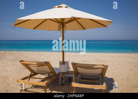 Due lettini in legno e ombrellone sulla spiaggia di sabbia con mare turchese. Vacanze senza persone sulle isole della Grecia Foto Stock