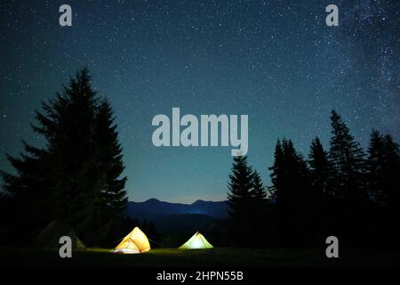 Luminose tende turistiche illuminate che brillano sul campeggio in montagne scure sotto il cielo notturno con stelle scintillanti. Concetto di stile di vita attivo. Foto Stock