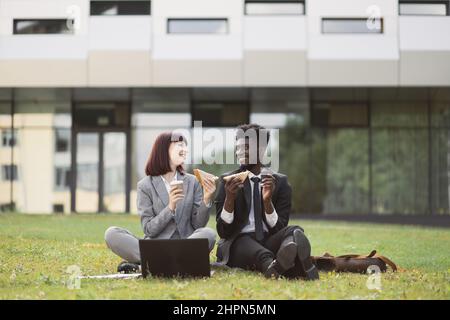 Stile di vita sano, pranzo al lavoro. Due colleghi multietnici, seduti sul prato fuori dall'ufficio, mangiando sandwich durante la pausa pranzo e parlando Foto Stock