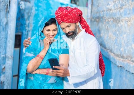 Felice coppia indiana tradizionale utilizzando lo smart phone, uomo sorridente indossando rosso turbante e donna in sari con telefono cellulare. Rajasthani persone che usano technol Foto Stock