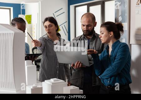 Coppia di architetti professionisti che tengono un notebook che guardano i progetti accanto ai colleghi con un tablet che punta a 3D maquette di grattacielo. Team di ingegneri architettonici che fanno lavoro di squadra creativo . Foto Stock