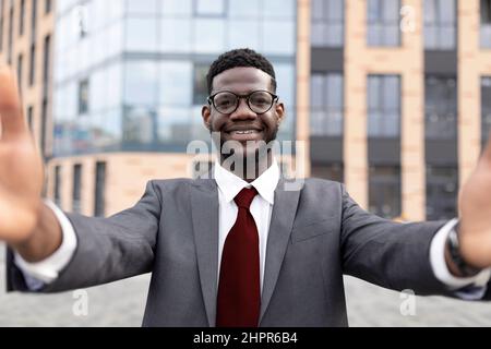 Un felice uomo d'affari afro-americano in costume formale che prende selfie mentre si trova fuori dal centro affari, catturando momento Foto Stock