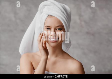 Concetto di pelle perfetto e salutare. Felice bella signora avvolto in asciugamano, applicando la crema sul viso e sorridendo alla macchina fotografica Foto Stock