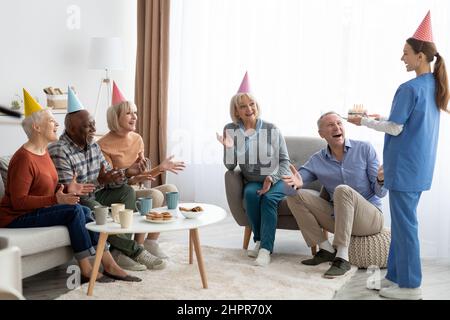 Infermiera amichevole che tiene torta di compleanno, persone anziane che hanno partito Foto Stock