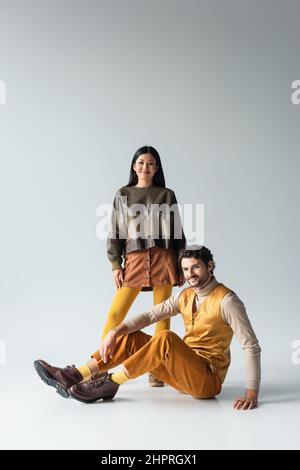 uomo sorridente in pantaloni gialli seduti vicino a donna asiatica in giacca di pelle grigio Foto Stock