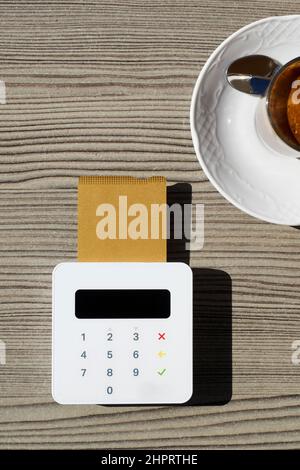 vista ad alto angolo del terminale di pagamento wireless, con una carta di credito inserita in esso, accanto a una tazza di caffè sul tavolo di un caffè sul marciapiede Foto Stock