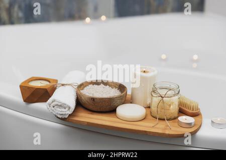 Preparazione per il trattamento spa dell'hotel, procedura per il bagno in casa. Lavabo bianco in bagno, accessori su vassoio candele Burning, sapone, spazzola per il piede, asciugamano, bottiglia di vetro con sale marino, fiore di orchidea Foto Stock