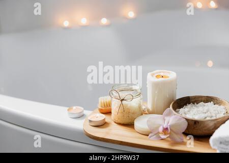 Preparazione per il trattamento spa dell'hotel o per la procedura di bagno in casa. Lavabo bianco in bagno con accessori su vassoio candele Burning, panno, sapone, spazzola per il piede, bottiglia con sale marino, fiore di orchidea Foto Stock