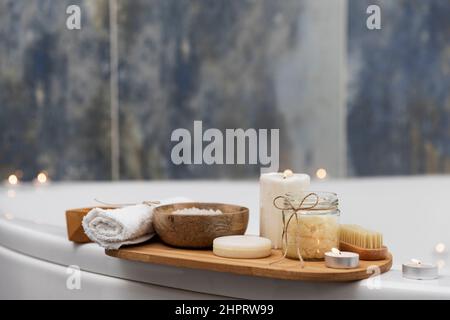 Preparazione per il trattamento spa dell'hotel, procedura per il bagno in casa. Lavabo bianco in bagno, accessori su vassoio candele Burning, sapone, spazzola per il piede, asciugamano, bottiglia di vetro con sale marino, fiore di orchidea Foto Stock