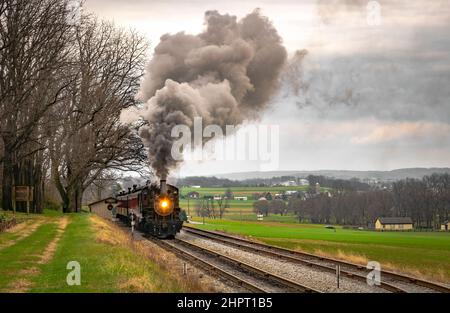 Un treno passeggeri a vapore antico che viaggia attraverso le fattorie Puffing un sacco di fumo in un giorno invernale nuvoloso Foto Stock