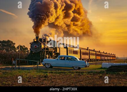 Una vista di un treno passeggeri a vapore antico che si avvicina all'alba con una testa piena di vapore e fumo che viaggia attraverso le terre con un'auto d'epoca in attesa di passare Foto Stock