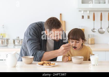 Concetto di cura dei bambini. Giovane padre amante che mangia il figlio piccolo con cereali a casa, mangiando la colazione insieme in cucina Foto Stock