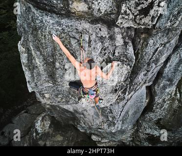 Giovane arrampicatore arrampicarsi su un percorso difficile su un masso alto con corda. Arrampicata su roccia senza paura durante il giorno d'estate. Concetto di sport estremo, avventure e stile di vita attivo. Vista aerea Foto Stock