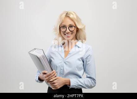 Amichevole giovane donna d'affari che tiene cartella con rapporti di affari in mani, sorridendo alla macchina fotografica su sfondo chiaro Foto Stock