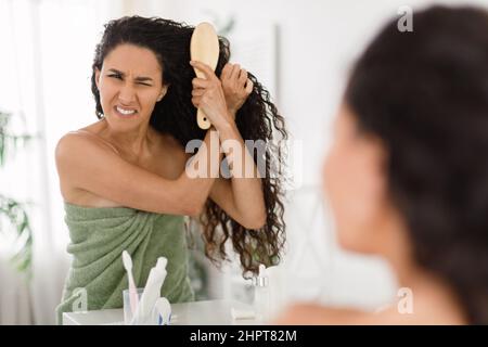 Frustrata giovane signora che ha problema spazzolando i suoi capelli aggrovigliati di fronte allo specchio a casa, spazio copia Foto Stock