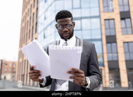 Giovane uomo nero in vestito che controlla i rapporti o che si prepara per il colloquio di lavoro, tenendo documenti o CV, in piedi all'aperto Foto Stock