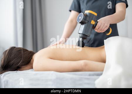 La donna massaggia la schiena del paziente con un massaggiatore a percussione Foto Stock