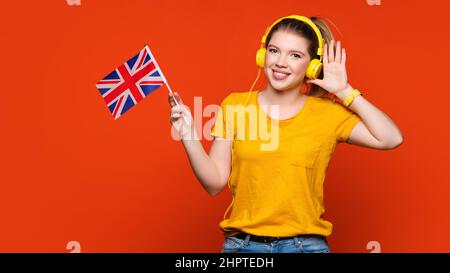 Ragazza in tuta gialla di base e cuffie tiene piccola bandiera inglese. Studiare all'estero e viaggiare. Programma internazionale di scambio studentesco. Imparare l'inglese britannico con nativo. Lingua straniera per orecchio Foto Stock