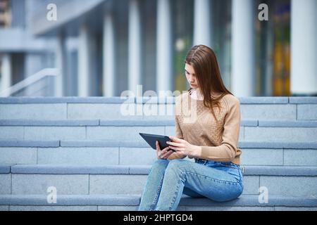 adolescente femminile si siede sui gradini, usa un tablet. giorno estivo soleggiato. copyspace. Foto Stock