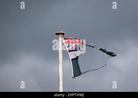 Una bandiera Union Jack strappata vola da un flagpole bianco in un giorno di luna. Le nuvole scure si aggiungono all'immagine scura. Foto Stock