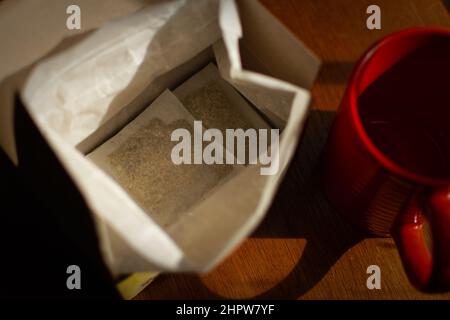 Una scatola aperta di bustine di tè e tazza rossa si siede su un tavolo di legno in luce d'ora d'oro. Foto Stock