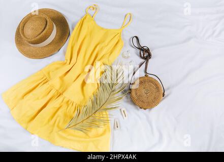 Cappello di paglia, abito giallo, borsa di paglia su sfondo bianco. Outfit estivo alla moda. Stay piatto con vestiti per vacanze e vacanze. Foto Stock