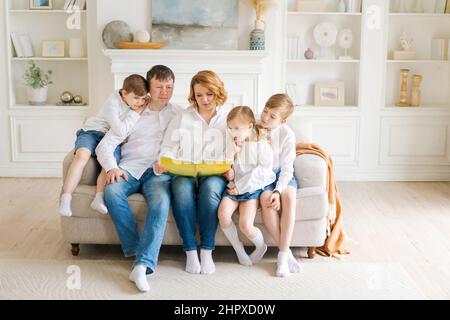 Buon libro di lettura della famiglia a casa. Mamma, papà e bambini si siedono insieme su un comodo divano in un nuovo grande e luminoso soggiorno con pareti bianche, mobili e ripiani sullo sfondo. Foto Stock