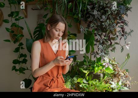 Casalinga caucasica seduta sul tavolo contro parete di fondo con piante interne nel giardino di casa e SMS utilizzando il telefono e Internet in una giornata di sole in un abito arancione Foto Stock